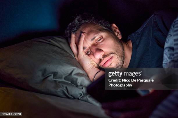 man lying awake in bed using smartphone at night - chronic illness stock pictures, royalty-free photos & images
