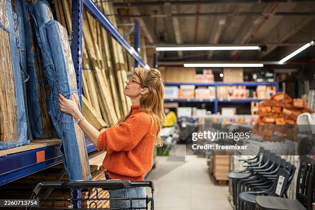 woman in orange sweater browsing lumber and boards in a busy hardware store aisle - hardware store stock pictures, royalty-free photos & images