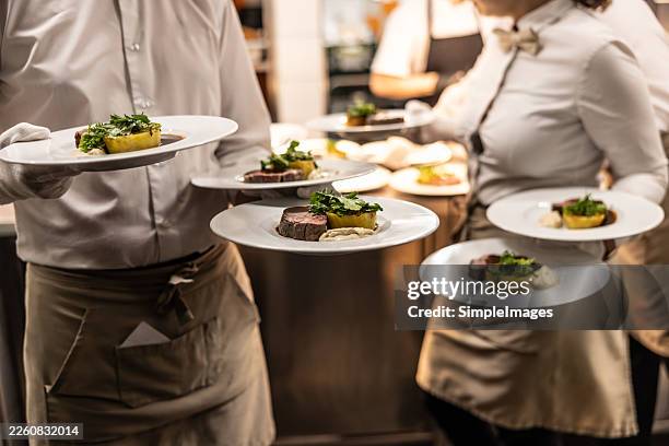 a close-up of waiters in white uniforms professionally carrying several plates of elegantly presented food. - restaurant work stock pictures, royalty-free photos & images
