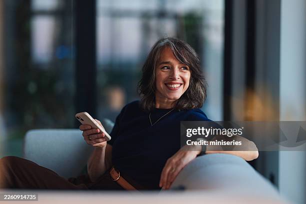 smiling senior businesswoman relaxing on a couch with smartphone in modern office lounge - chairperson stock pictures, royalty-free photos & images