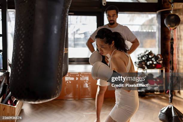 female boxer practicing strikes on heavy bag with male coach in gym - mixed martial arts stock pictures, royalty-free photos & images