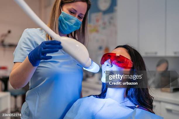 young female patient getting her teeth whitened at the dentist's office by a mid-adult female dentist - tooth whitening stock pictures, royalty-free photos & images