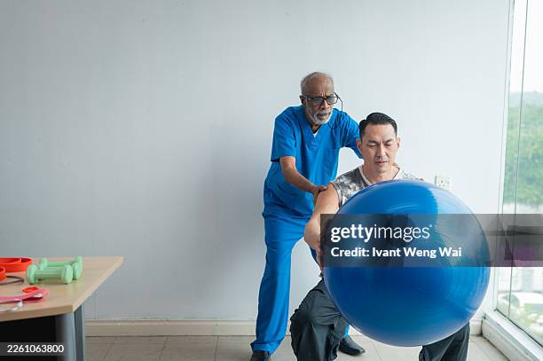 hombre asiático de media edad haciendo ejercicio con un fisioterapeuta usando práctica de pelota de fitness en una clínica médica foto de archivo - achatar la curva de epidemia fotografías e imágenes de stock