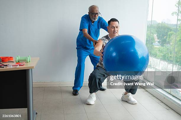 hombre asiático de media edad haciendo ejercicio con un fisioterapeuta usando pelota de fitness en una clínica médica foto de stock - achatar la curva de epidemia fotografías e imágenes de stock