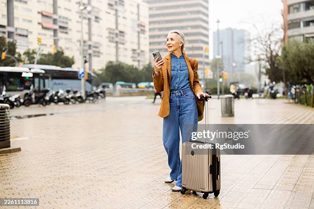 mature woman traveling, pulling suitcase and holding smartphone in city - solo travel stock pictures, royalty-free photos & images