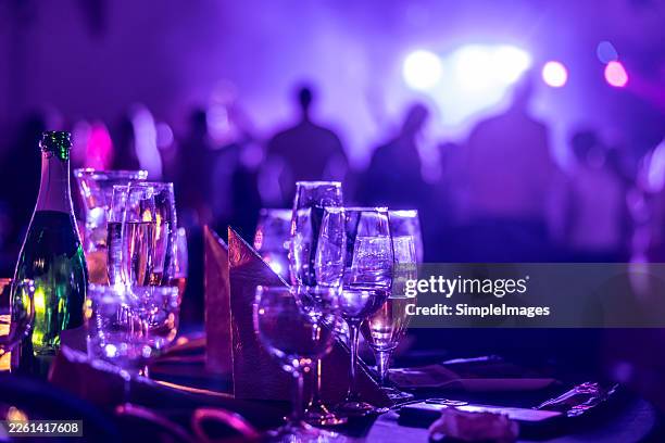a table full of glasses and a bottle of sparkling wine dominates the foreground in atmospheric purple lighting. in the blurred background, figures of people on the dance floor are outlined, which completes the dynamic mood of the ongoing celebration. - locale notturno foto e immagini stock