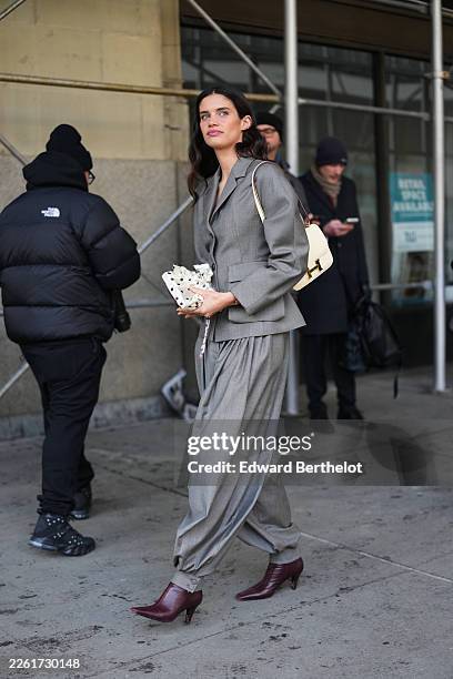 Sara Sampaio wears loose wavy dark brown hair, a white leather Constance shoulder bag from Hermes, a gray wool puffy sleeves / shoulder pads / belted...