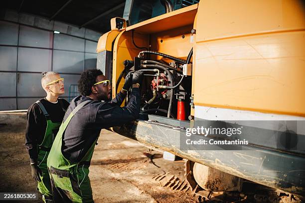 two mechanics in safety glasses and overalls inspect the engine of a large yellow construction vehicle - repair shop stock pictures, royalty-free photos & images