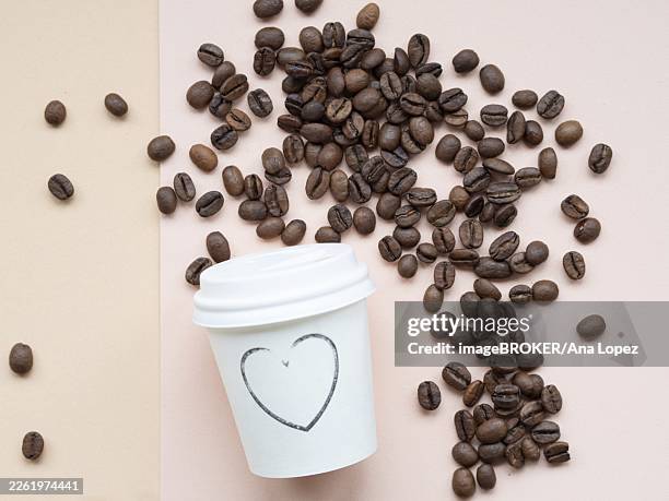roasted coffee beans scattered on a two-tone background near a paper cup with a heart drawn on it, conveying a love for coffee - caffeine stock pictures, royalty-free photos & images