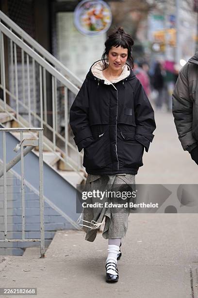 Guest wears dark brown hair in a bun with bangs, a black hooded winter jacket "LIT active" with a cream hood lining, a gray plaid skirt, white socks,...