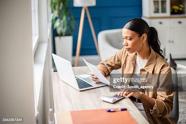 young woman works on her taxes at a home office desk while using a calculator and laptop - financial literacy stock pictures, royalty-free photos & images