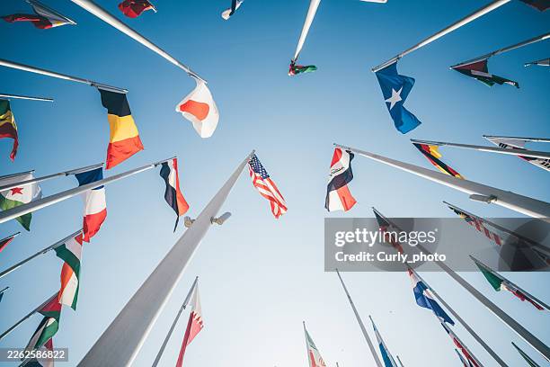 a low-angle view of diverse international flags waving on tall poles against a clear blue sky. this vibrant display symbolizes global unity, international relations, and the spirit of cooperation between nations. - diplomatie stock-fotos und bilder