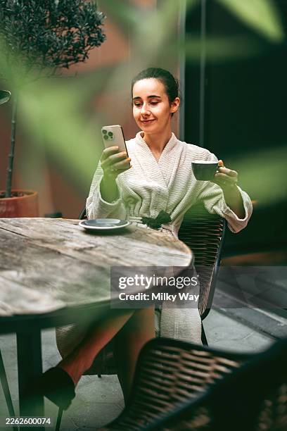 young woman sitting at terrace table holding smartphone and espresso cup. green leaves foreground blur. concept of remote communication, lazy home weekend, relaxed urban lifestyle, digital nomad mood and coffee break connection. - facilité photos et images de collection