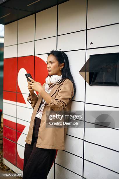 young woman using smartphone at parcel locker with headphones while waiting to pick up a package - curbside pick stock pictures, royalty-free photos & images
