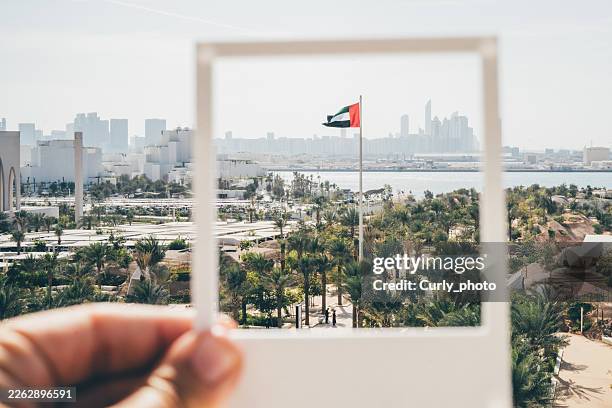 a hand holds a white frame framing the uae flag against a backdrop of a modern city skyline and palm trees. - abu dhabi skyline stock pictures, royalty-free photos & images