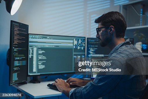 Young Latin male software developer working late analyzing information and code on multiple displays in office at night. Artificial intelligence, machine learning and data innovation in business. Young Latin male software developer working late analyzing information and code on multiple displays in office at night. Artificial intelligence, machine learning and data innovation in business.