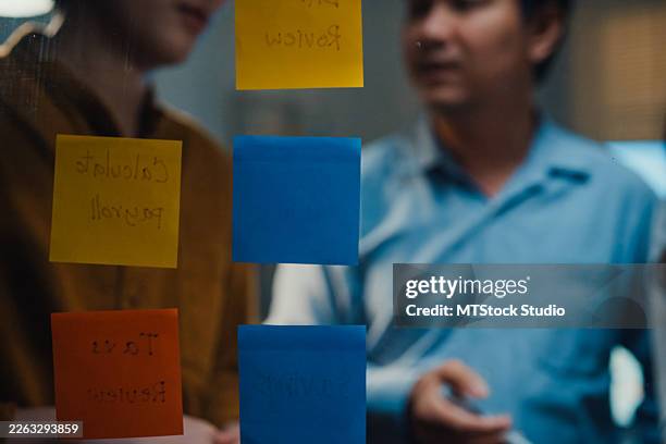 close-up of asian business team meeting brainstorms using sticky notes on a glass wall to map ideas and prioritize next steps for a project in office at night. - marktonderzoek stockfoto's en -beelden