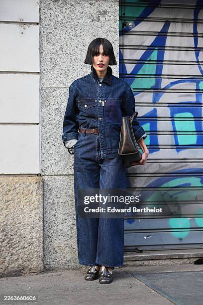 Lisha Ho wears black bobbed hair with bangs, a blue dark denim jacket with a black dark corduroy collar, blue dark denim wide leg jeans, a brown...