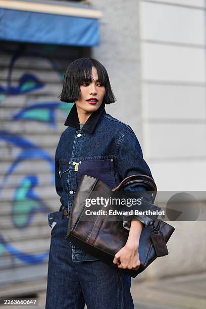 Lisha Ho wears black bobbed hair with bangs, a blue dark denim jacket with a black dark corduroy collar, blue dark denim wide leg jeans, a brown...