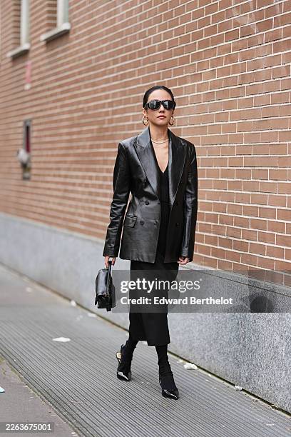 Yoyo Cao wears black hair in a bun, black futurist sunglasses, gold large earrings, a black varnished leather double breasted blazer, a black V-neck...