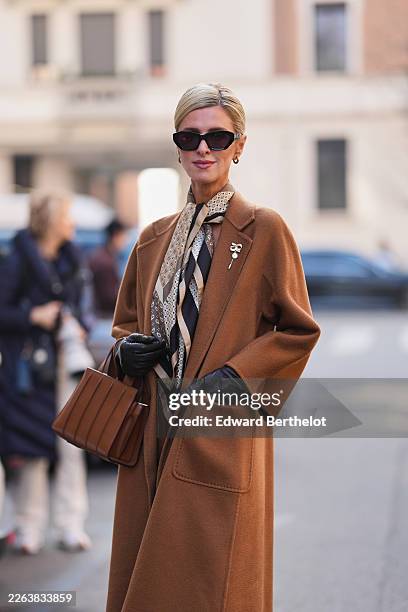 Nicky Hilton Rothschild wears blonde hair in a bun, black sunglasses, gold earrings, a brown wool relaxed coat with gold brooch, a beige / brown /...