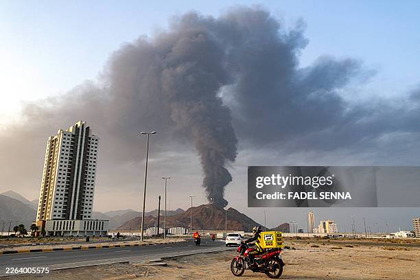 Delivery-persons ride motorcycles along a road as behind a tall smoke plume billows following an explosion in the Fujairah industrial zone on March...