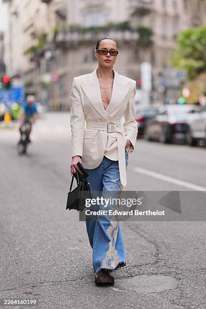 Guest wears brown hair in a ponytail, brown tinted sunglasses, a cream tailored belted blazer jacket, blue denim wide leg jeans with embroidered...