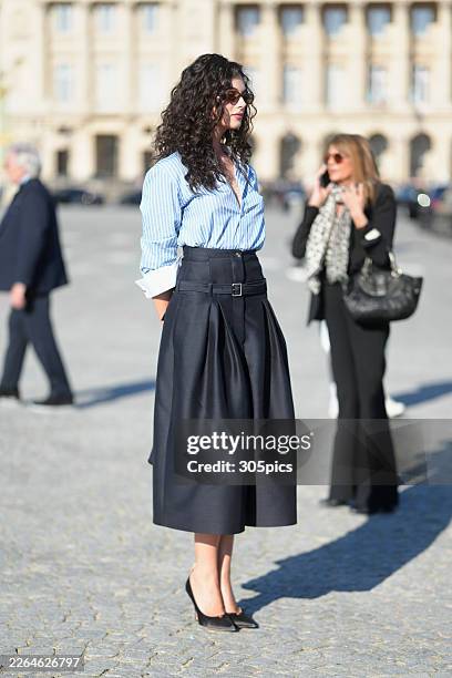 Deva Cassel is seen wearing brown tortoiseshell sunglasses, a Dior blue and white striped button-down shirt with white cuffs, a black high-waisted...