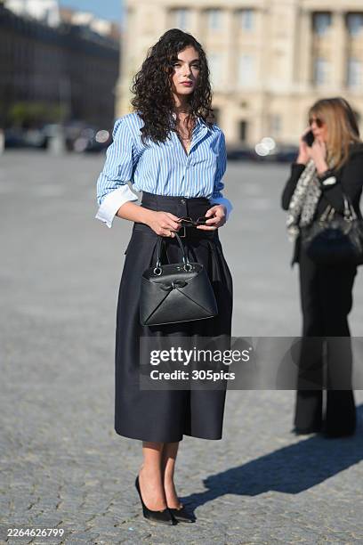 Deva Cassel is seen wearing brown tortoiseshell sunglasses, a Dior blue and white striped button-down shirt with white cuffs, a black high-waisted...