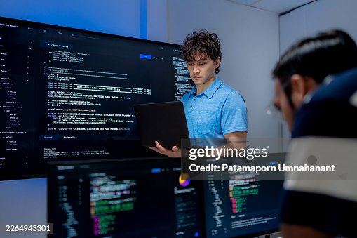 Software developer reviewing source code on a large monitor while a colleague updates system firmware via coding. IT professionals collaborating on programming and maintenance in a modern server room office. Software developer reviewing source code on a large monitor while a colleague updates system firmware via coding. IT professionals collaborating on programming and maintenance in a modern server room office.