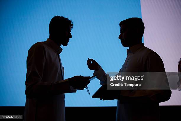 two men standing facing each other, sharing a digital tablet. two men in silhouette are standing facing each other, sharing a digital tablet. - arab tablet stock pictures, royalty-free photos & images