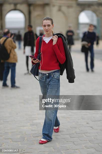 Sadie Pickup is seen wearing a white t-shirt, a red ribbed V-neck sweater, blue baggy denim jeans, white socks, and red leather ballet flats, while...