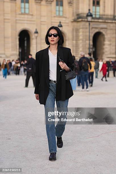 Guest wears black hair in a bob haircut, black sunglasses, a black tailored blazer, a white cotton T-shirt, blue denim straight leg jeans, a black...