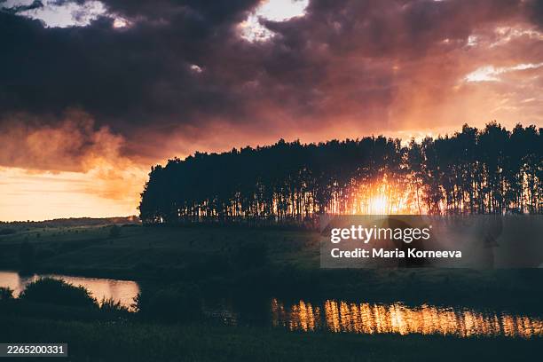 a dramatic sunset glows behind a silhouette of trees reflecting in a calm river under a stormy sky. - cielo romantico foto e immagini stock