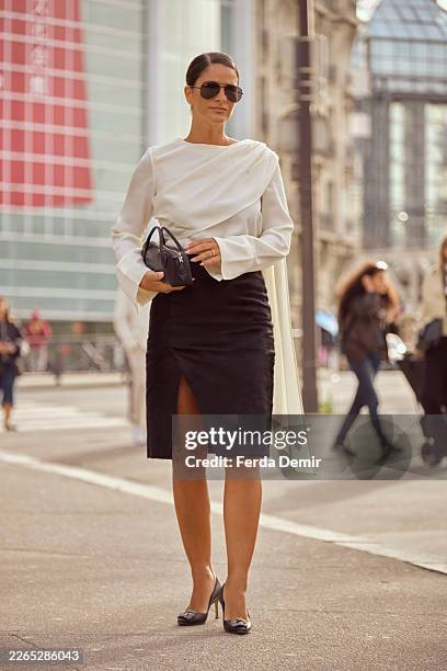 Gili Biegun wears a white draped asymmetric blouse with wide flared sleeve paired with a black high-waisted pencil skirt with front slit, aviator...