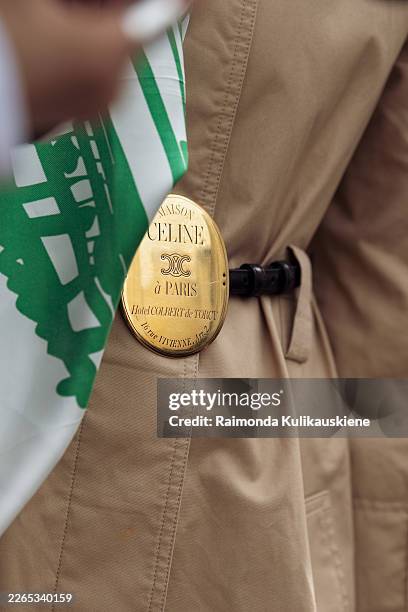 Guest wears a beige trench coat and a black belt with golden Celine buckle, a white scarf with large green graphic print draped over the shoulders...