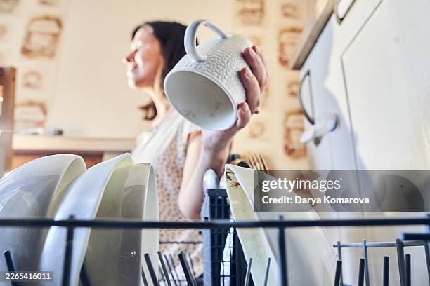 woman unloading clean dishes from dishwasher in cozy kitchen while holding a ceramic mug - unloading dishwasher stock pictures, royalty-free photos & images