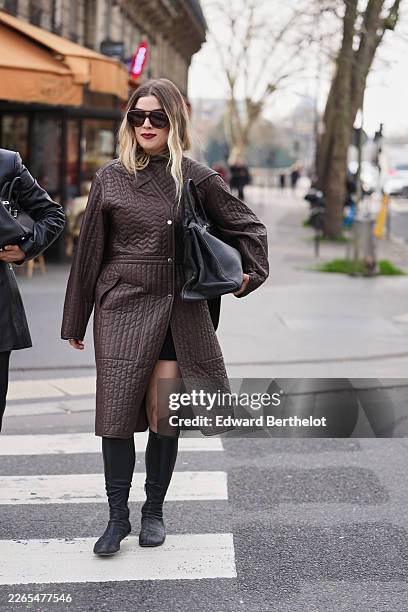 Guest wears dark brown oversized aviator sunglasses, a dark brown quilted leather long coat, a black mini skirt, a black leather oversized Birkin...