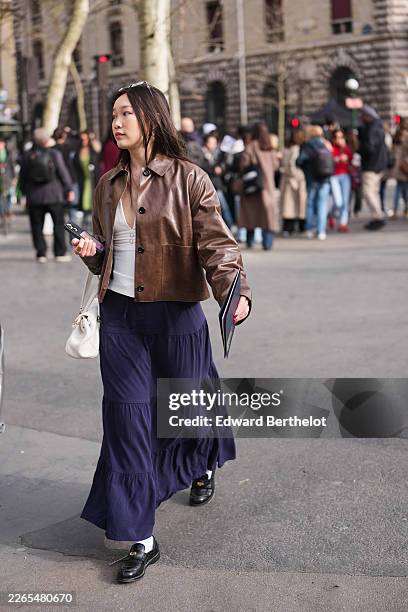 Guest wears dark brown hair, a brown leather jacket, a white ribbed V-neck fitted top, a dark blue long skirt, a white leather bag, black leather...