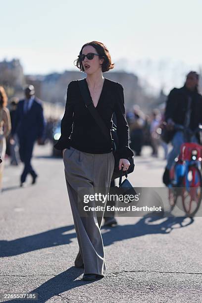 Guest wears dark brown bob haircut, black sunglasses, a black V-neck / long sleeves top, gray woven wide leg trousers, a black leather crossbody...