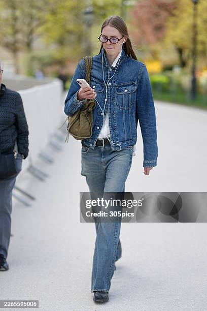 Person is seen wearing burgundy oval glasses, white wired earphones, a blue denim jacket, a white collared shirt, a black leather belt with a silver...