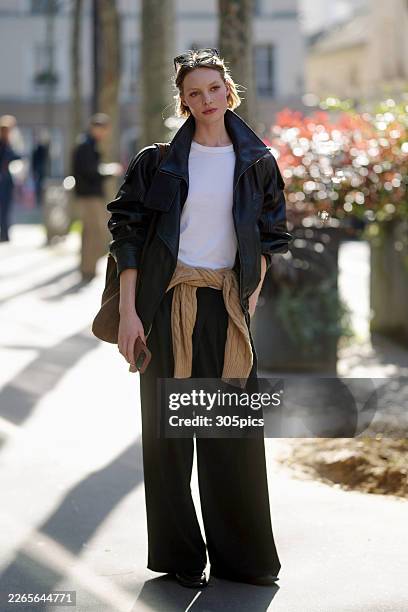 Maya Vozhdaeva is seen wearing brown tortoiseshell sunglasses, a white crewneck t-shirt, a black oversized leather jacket, a tan cable-knit sweater...