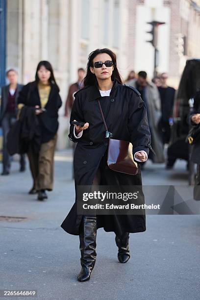 Guest wears dark brown hair, sunglasses, a black cotton trench coat, a white cotton shirt, black pants, a dark brown varnished leather bag, black...