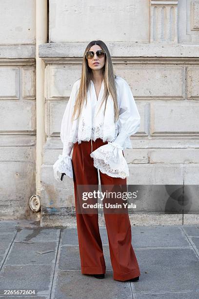 Guest wears a romantic white blouse with dramatic lace-trimmed cuffs and hem paired with high-waisted rust wide-leg trousers, oversized gradient...