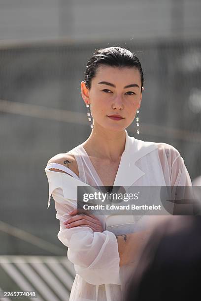 Amalie Gassmann wears white pearls pendant earrings, black hair in a bun, a white sheer chiffon loose coat, white cotton fitted long dress, outside...