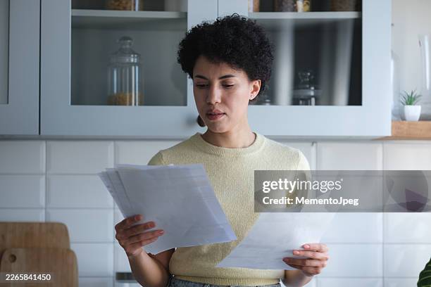 upset young african american woman holding utility bills feeling frustrated managing monthly household budget, standing in kitchen with financial papers in hands - struggle stock pictures, royalty-free photos & images