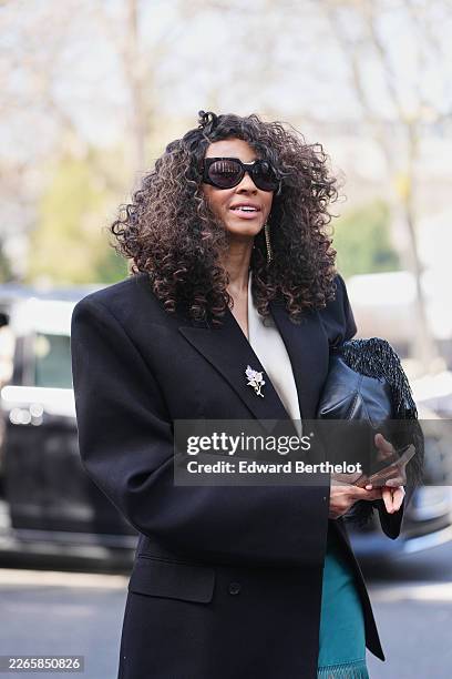 Guest wears brown curly hair, black sunglasses, a black wool oversized blazer with silver brooch, a cream V-neck blouse top, a blue green satin /...