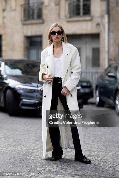 Guest wears silver hoop earrings, black sunglasses, a white v-neck t-shirt, a white long coat, a black leather belt, a black leather bag, black...