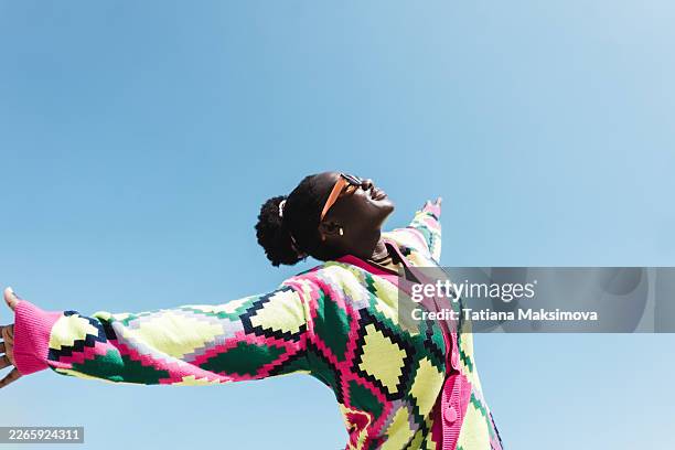 a young black woman with joyful expression tilts her head back laughing, wearing colorful knitted sweater and retro sunglasses against bright blue sky. - eccentric stock pictures, royalty-free photos & images