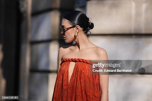 Guest wears black hair in a bun, brown yellow tinted sunglasses, a red sheer lace strapless / cut-out chest / balloon loose dress, outside...